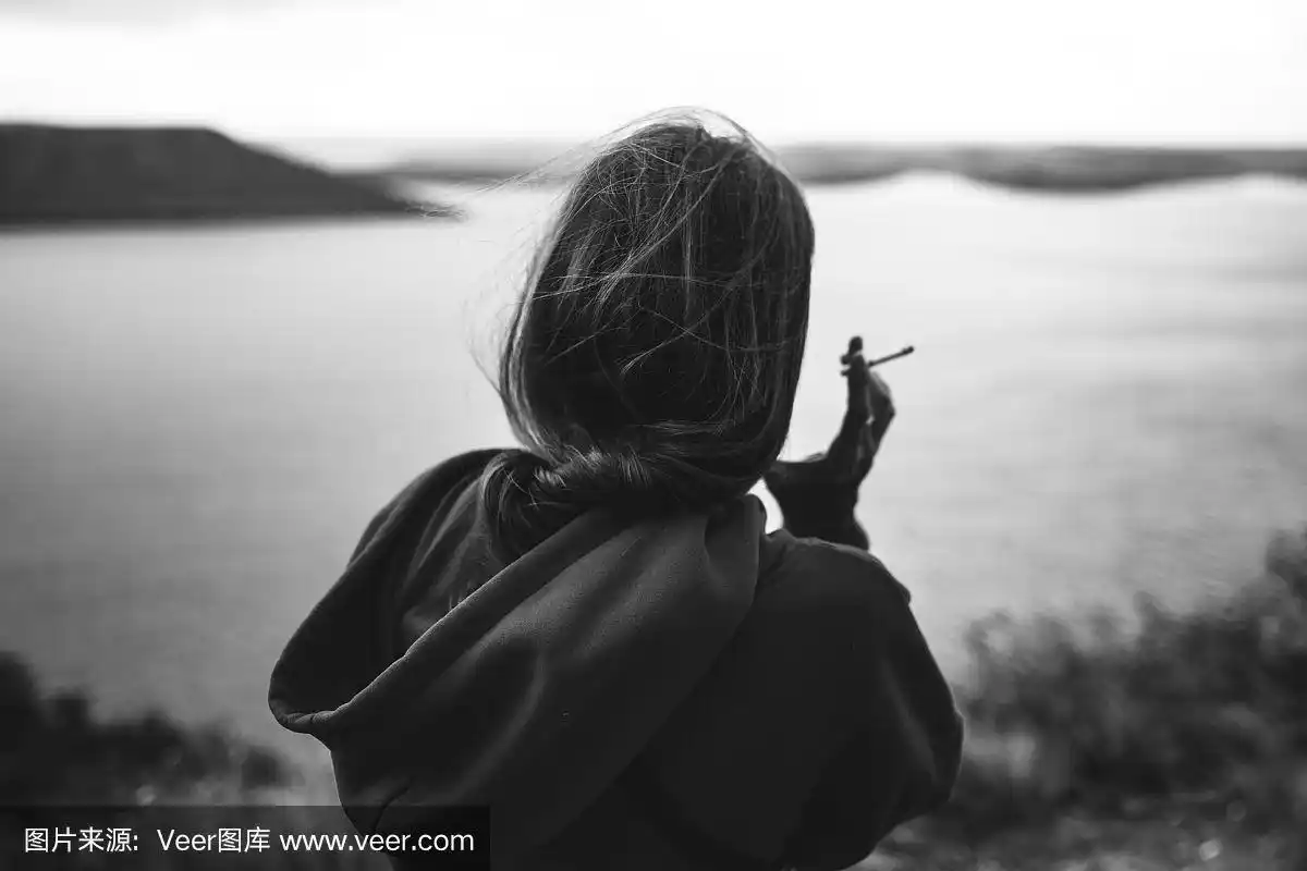 穿着风衣,手拿香烟,站在岩石山顶上看河的时髦女孩的背影.