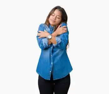 年轻漂亮的黑发女人穿着蓝色牛仔衬衫,在孤立的背景下拥抱自己,快乐而